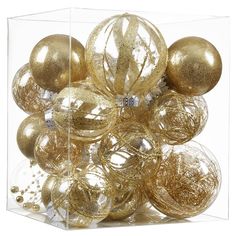 a clear box filled with gold ornaments on top of a white surface and surrounded by smaller shiny balls