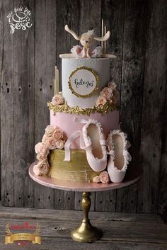 a pink and gold cake with ballet shoes on top is sitting on a stand in front of a wooden wall