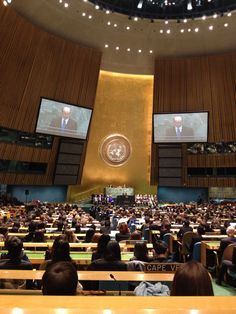 a large room filled with people sitting at desks in front of two screens on the wall