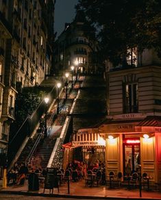 people are sitting at tables on the steps in front of buildings and stairs lit up by street lights