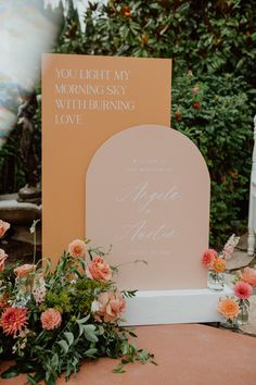 an orange and pink wedding sign with flowers on the ground in front of some bushes