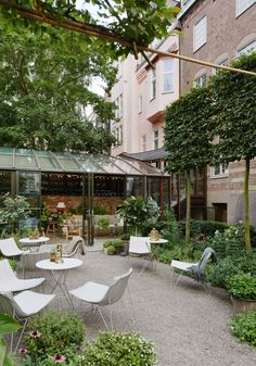 an outdoor seating area with white chairs and tables surrounded by greenery in front of a brick building