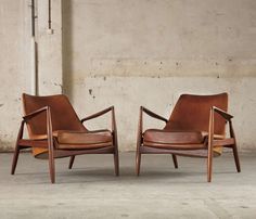 two brown leather chairs sitting next to each other on cement floored area with concrete wall in background