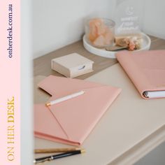 pink notebooks and pens on a desk with a white box in the corner behind them