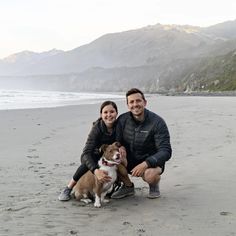 a man and woman pose with their dog on the beach in front of some mountains