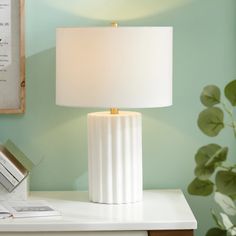 a white table lamp sitting on top of a desk next to a framed photograph and a plant