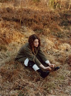 a woman sitting on the ground with her legs crossed and wearing a coat over her shoulders