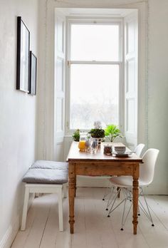 a dining table with two chairs and a bench in front of a window that has potted plants on it