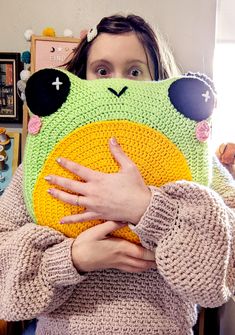 a woman holding a crocheted frog pillow in front of her face