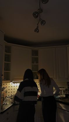 two people standing in a kitchen with lights on above the counter and below the cabinets