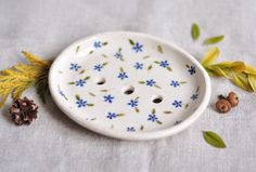 a white ceramic dish with blue flowers and leaves on the table next to some pine cones