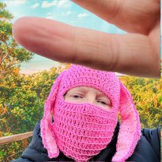 a woman wearing a pink crochet bunny hat and scarf is holding up her hand to the camera