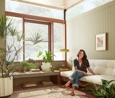 a woman sitting on a white couch in a living room next to a plant filled window