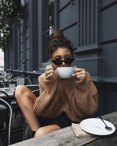 a woman sitting at an outdoor table drinking from a coffee cup with her eyes closed