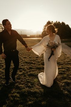 a bride and groom hold hands as the sun sets behind them in an open field