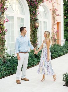 a man and woman are holding hands while walking down the sidewalk in front of some flowers