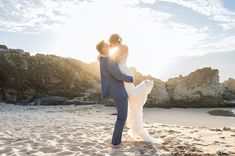 a bride and groom kissing on the beach in front of some rocks at sunset or sunrise