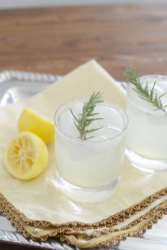 two glasses filled with lemonade and rosemary on a silver tray next to a slice of lemon