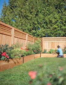 a woman sitting on the grass in front of a wooden fence and flower garden area