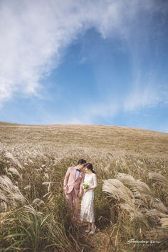 two women are standing in the middle of a field with tall grass and blue skies