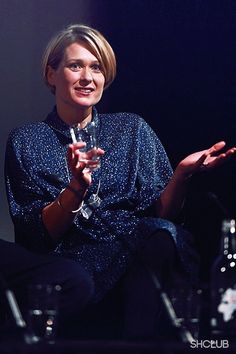 a woman sitting in a chair holding a wine glass and talking to someone on stage