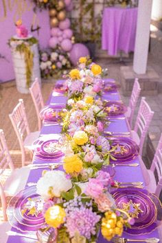 a long table is set with purple and yellow flowers, plates and napkins on it