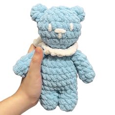 a hand holding a blue crocheted teddy bear in front of a white background