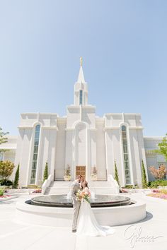 a bride and groom standing in front of a white church with flowers on the ground