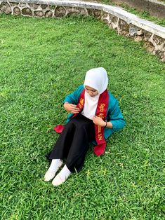 a woman sitting on the grass in front of a stone wall wearing a headscarf