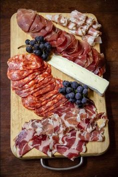 an assortment of meats and cheeses on a wooden cutting board with grapes, bread