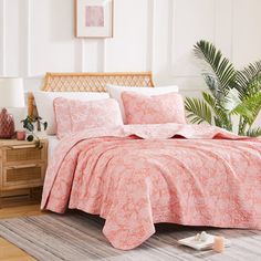 a bed with pink comforter and pillows in a room next to a plant on the floor