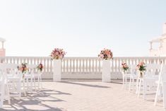 an outdoor ceremony setup with white chairs and floral centerpieces on the back wall