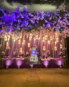 a wedding cake sitting on top of a table next to purple and white flowers hanging from the ceiling