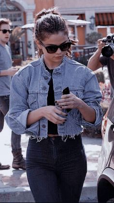 a woman in black shirt and jean jacket using her cell phone while standing next to a car