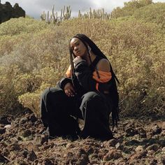 a woman with dreadlocks sitting on the ground in front of some cactus bushes