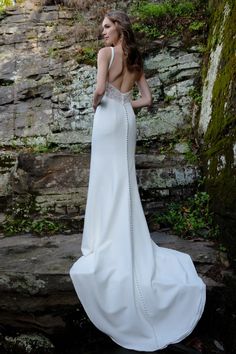 a woman in a white wedding dress standing near a stone wall with her back to the camera