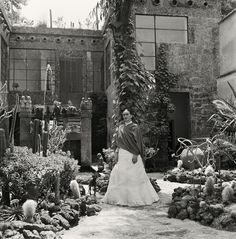 an old black and white photo of a woman standing in front of a house surrounded by plants