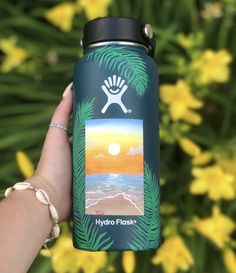 a person holding up a can of hydro flask in front of some yellow flowers