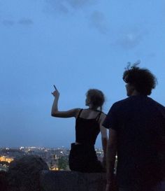 two people standing on top of a hill looking at the city lights in the distance