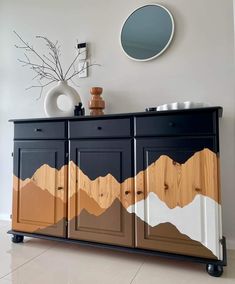 a dresser with mountains painted on it and a round mirror above the top, next to a vase