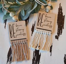 two wooden tags with words on them sitting next to some plants and greenery hanging from strings