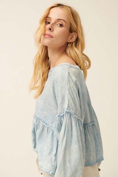 Acid washed denim blouse. Split neckline. 3-button closure with wood buttons. Long bubble sleeves with elastic cuffs. Drop shoulder. Exposed seams with frayed, raw-edge detailing. Flared ruffle hem. Babydoll silhouette. Loose fit. 100% Cotton. Imported. Designed in LA. Model wears size S. Exposed Seams, Wood Buttons, Acid Wash Denim, Denim Blouse, Babydoll Top, Washed Denim, Acid Wash, Raw Edge, Ruffle Hem