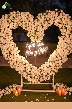 a heart - shaped sign with flowers and candles in front of it that says, will you marry me?