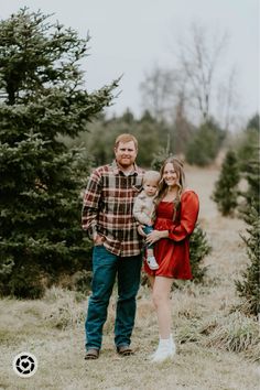 a man and woman holding a baby standing in front of christmas trees at the tree farm