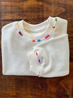 a white t - shirt with colorful beads is laying on a wooden surface and it's sleeves are rolled up