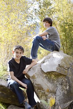 two boys are sitting on rocks in the woods, one is leaning against the rock