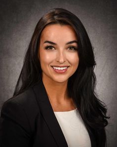 a woman with long black hair and a blazer smiles at the camera while standing in front of a dark background