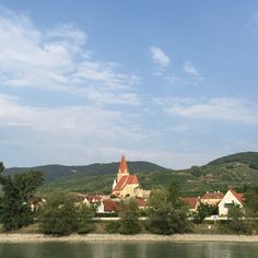 a church on the side of a river with mountains in the background and blue sky