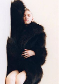 a woman in a black fur coat leaning against a wall