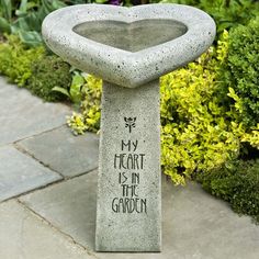 a cement heart shaped fountain sitting on top of a stone walkway next to green plants